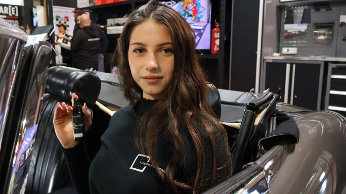 Young woman holding a camera, posing inside a car at an exhibition.