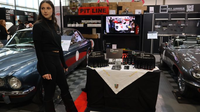 Donna in black attire stands beside a display table at a car exhibition with vintage cars.