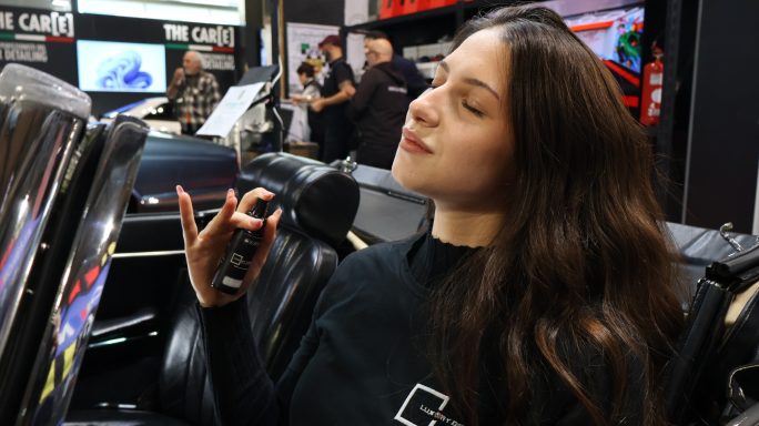 Young woman with long hair sitting in a vehicle, holding a microphone and smiling.
