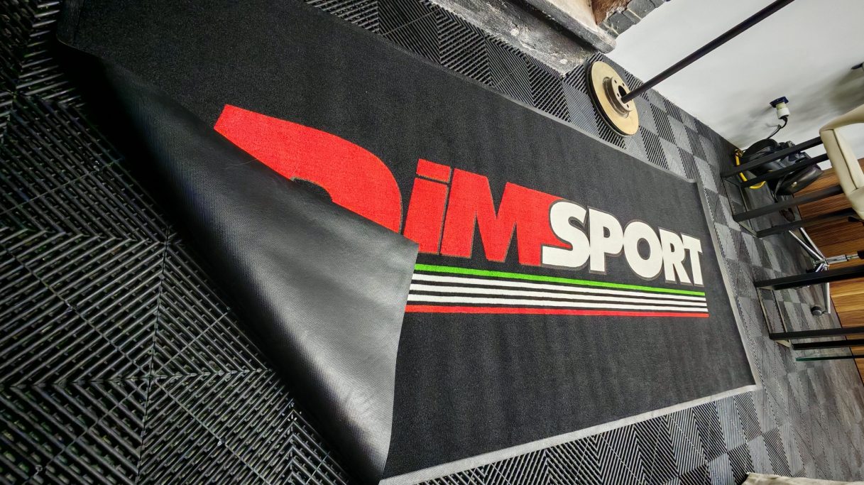 Tappeto nero con logo rosso "DIMSPORT" su sfondo grigio e strisce verdi.